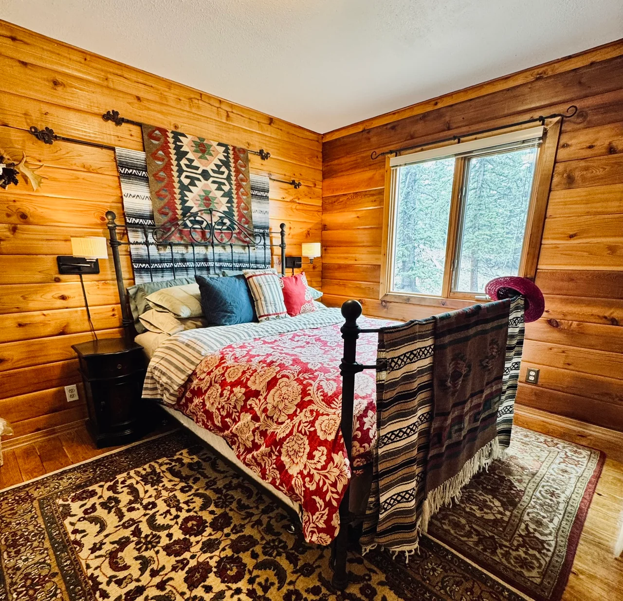 Dark metal bed with red patterned bedding, layered textiles and a tree-facing window in a wood-paneled room.