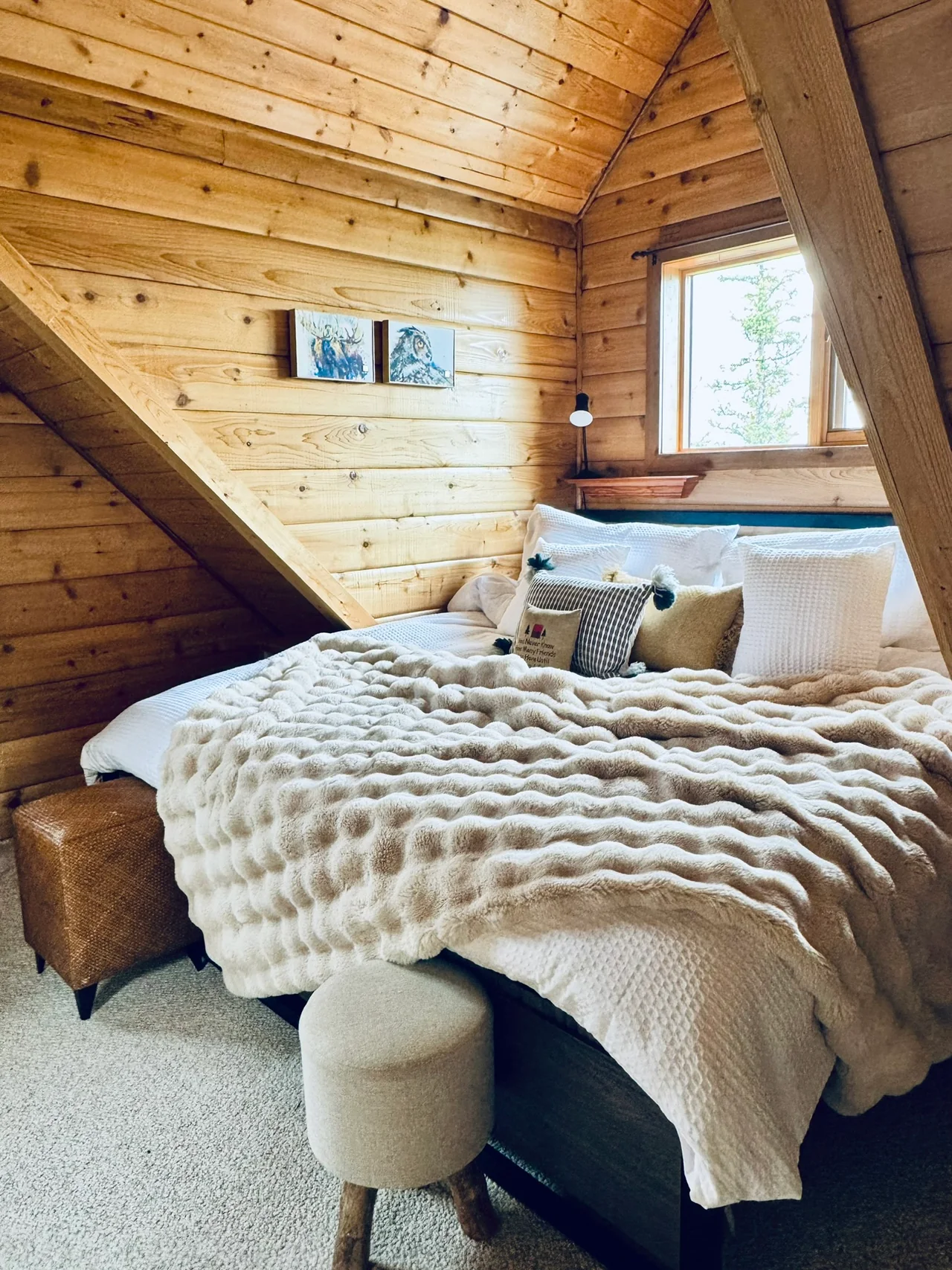 Bed with cream bedding and a textured throw below a window and sloping wood-paneled ceiling.
