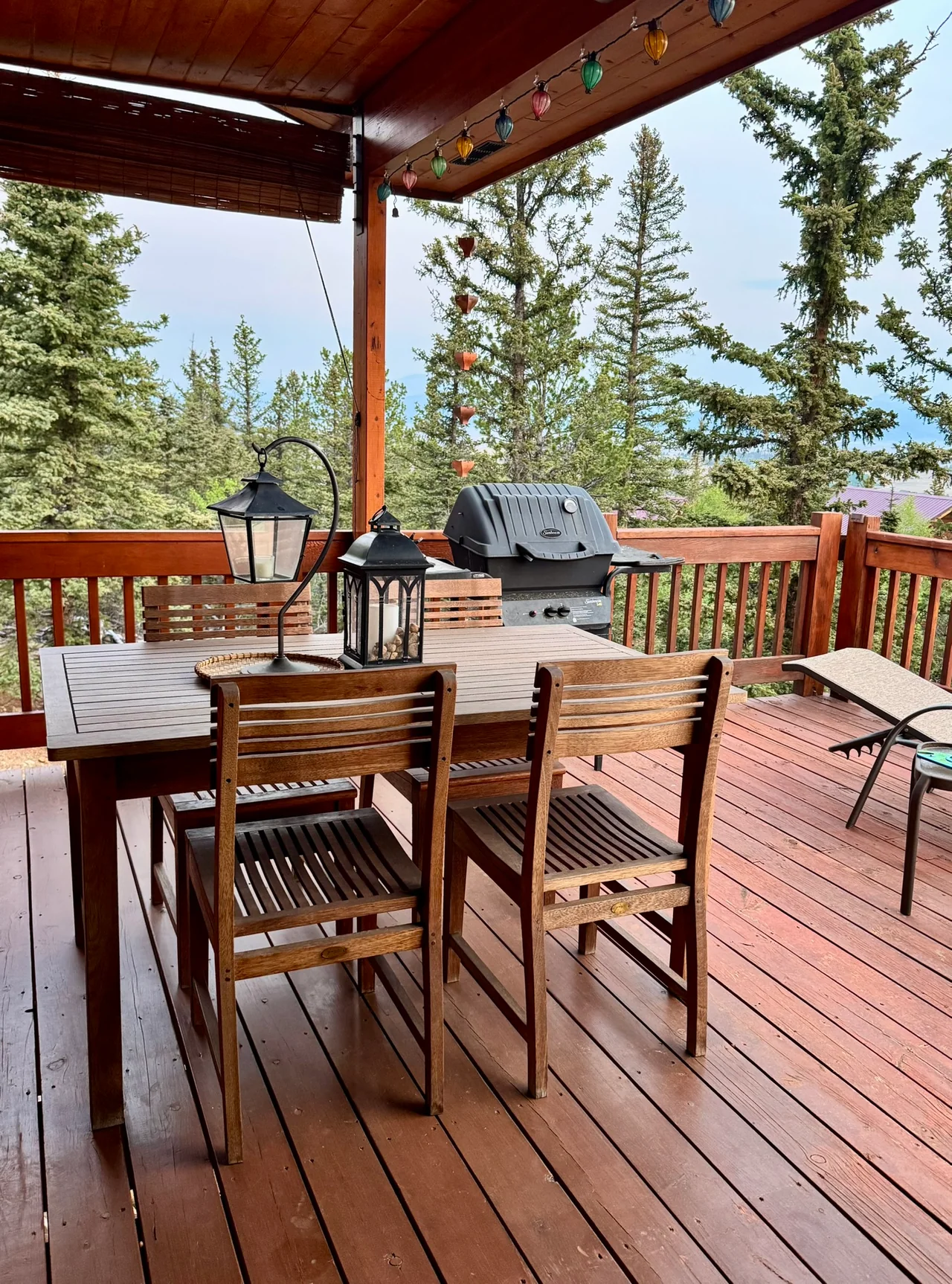 Wooden dining table, chairs and a grill on a covered deck edged by pine trees and hanging lights.