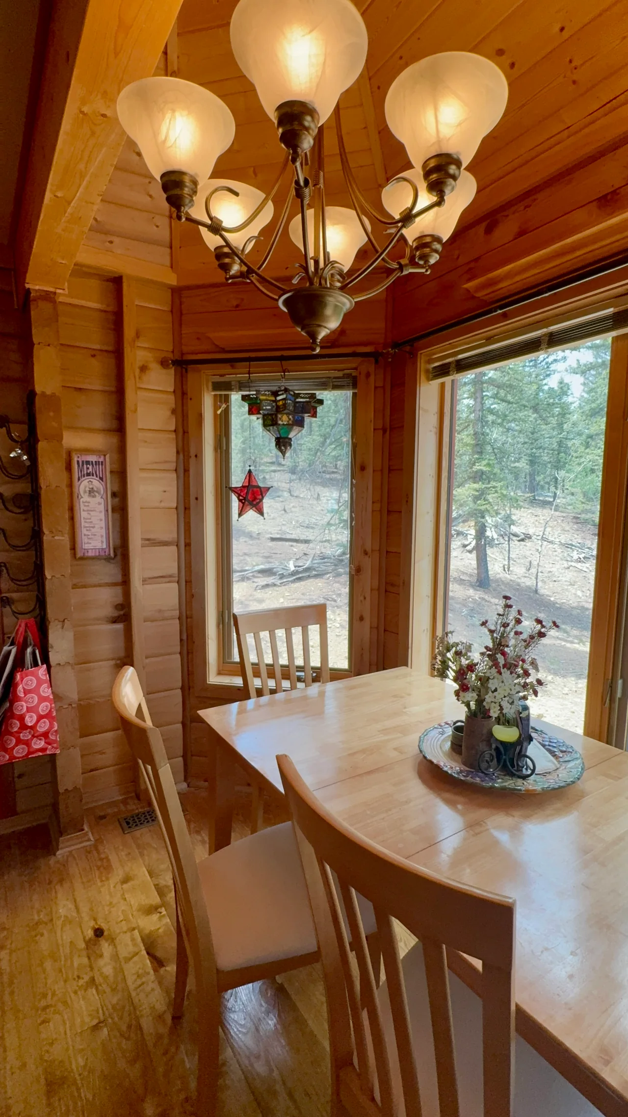 Wooden dining table and chairs beneath a chandelier, beside large windows facing the trees.