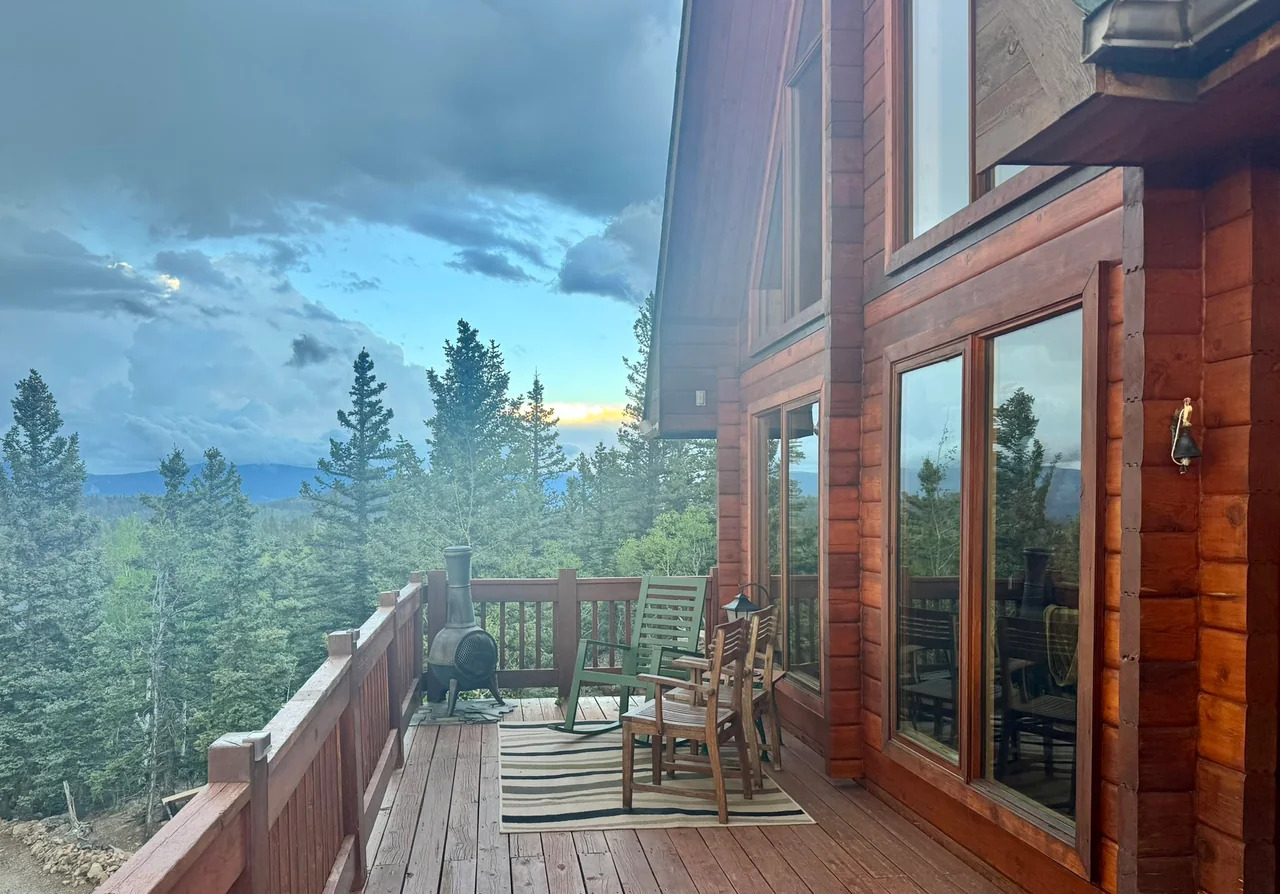 Wood-sided cabin and deck with two wooden chairs, pine trees and distant mountains beneath a cloudy sky.