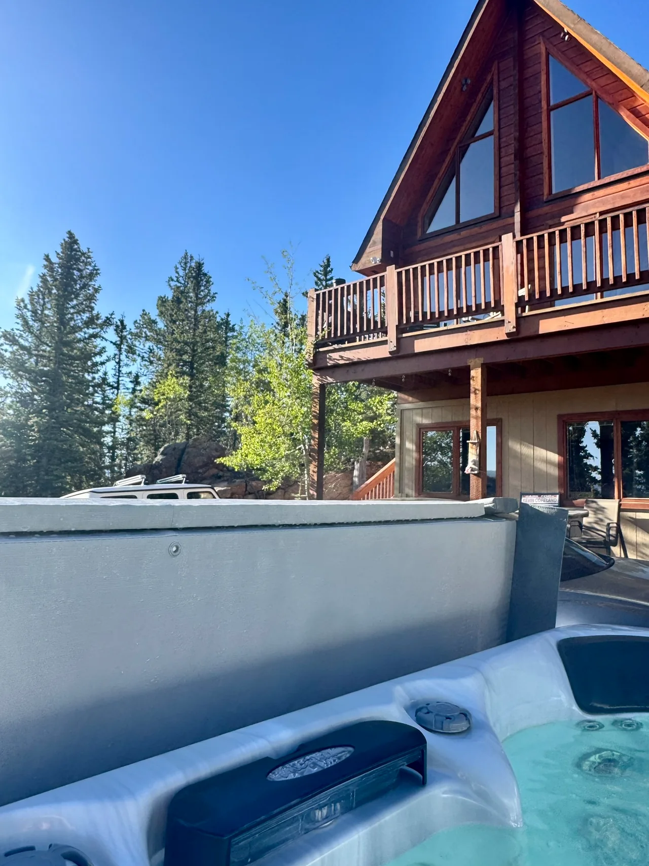 Water and seats in the hot tub, its raised cover and the cabin balcony beyond, with trees under a blue sky.
